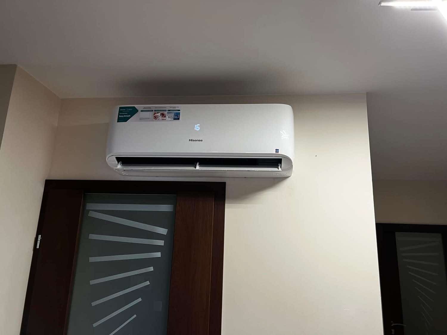 Biała klimatyzacja Hisense zamontowana nad ciemnymi drzwiami z geometrycznym wzorem na jasnej ścianie. Widoczny wyświetlacz temperatury.