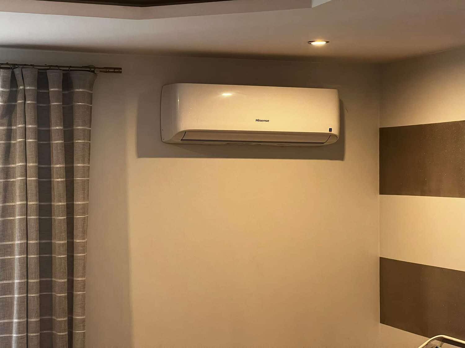 Biała klimatyzacja Hisense zamontowana na ścianie w pokoju z szarymi zasłonami i dekoracyjnymi pasami. Widoczne oświetlenie sufitowe.