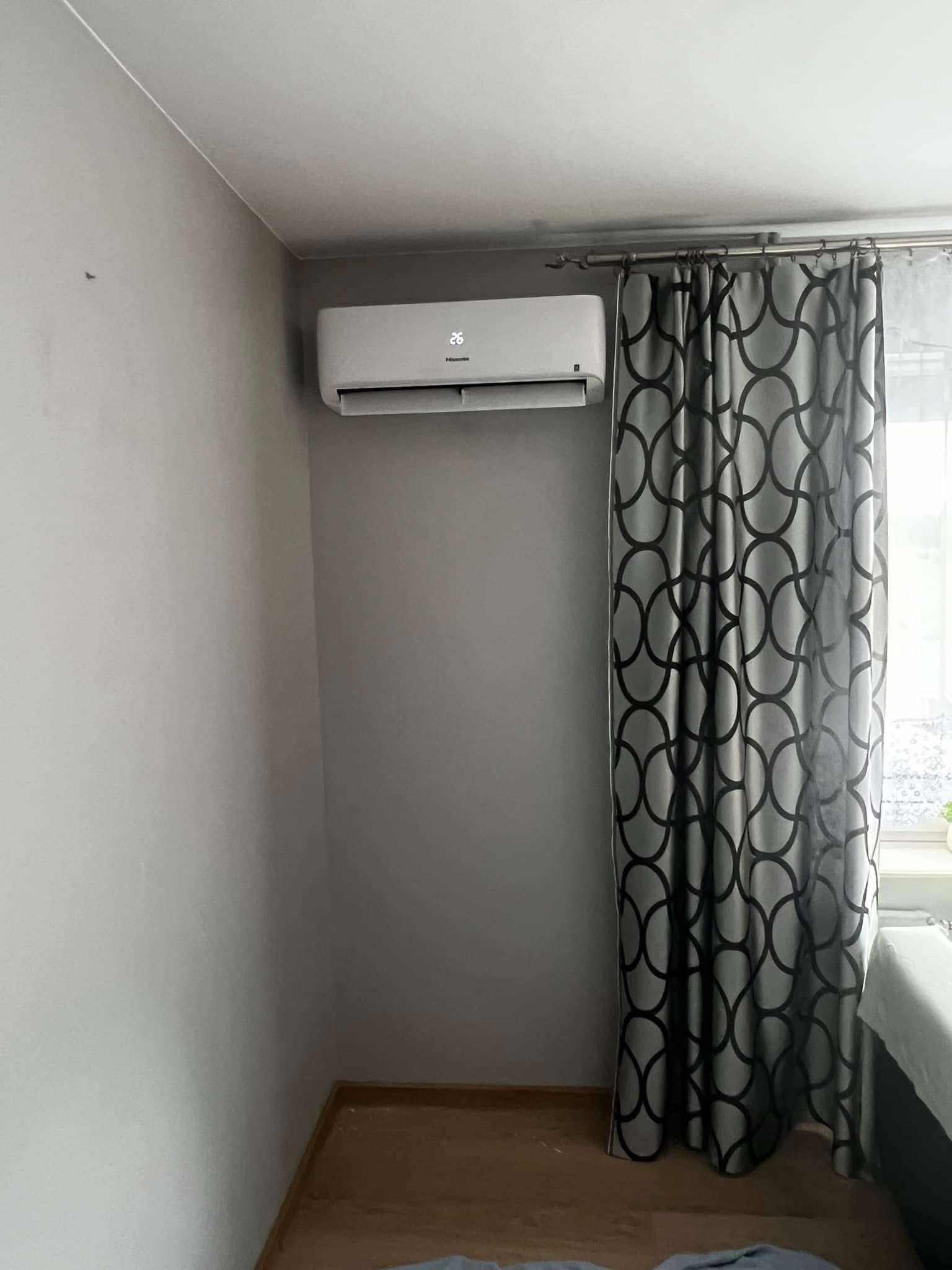 Biały klimatyzator ścienny Hisense, zamontowany w pokoju z szarą ścianą i wzorzystymi zasłonami. Widoczny wyświetlacz temperatury 26 stopni.