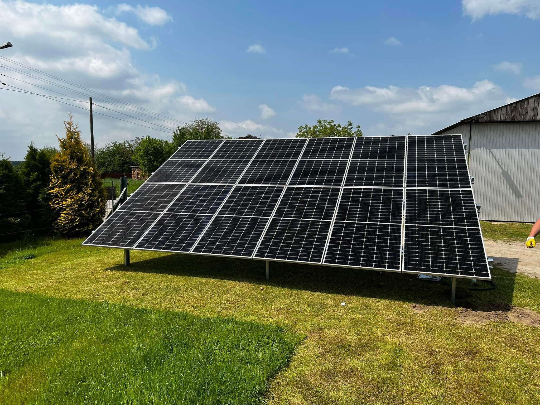 Panel fotowoltaiczny na trawniku, z widocznym budynkiem gospodarczym w tle. Instalacja składa się z kilkunastu modułów, zamontowanych na metalowej konstrukcji.