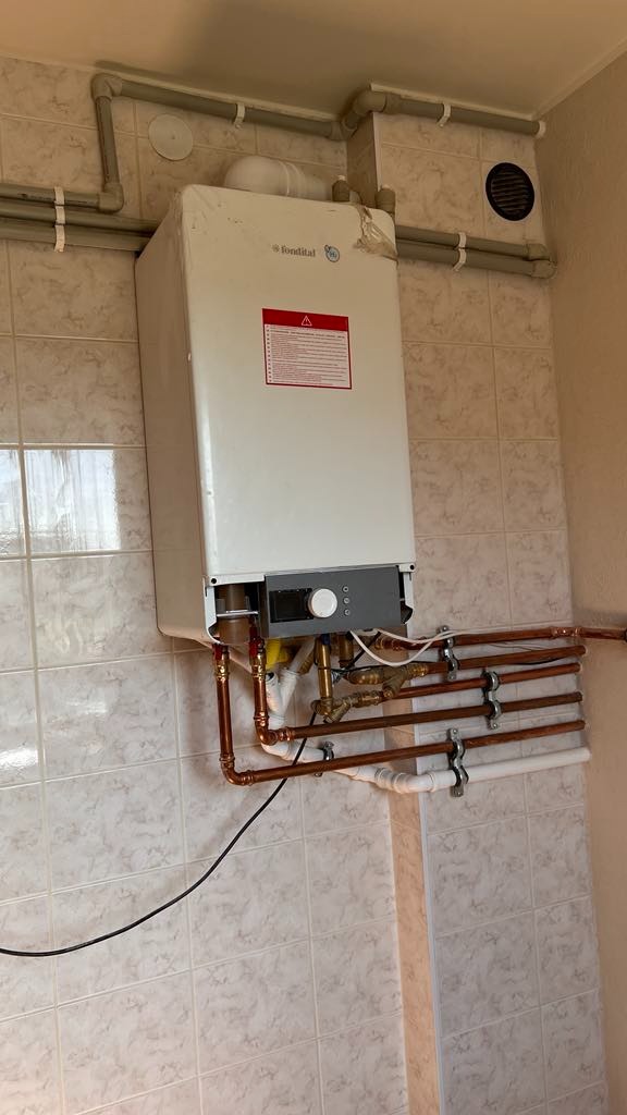 Biały piec gazowy Fondital zamontowany na ścianie z beżowych płytek, z widocznymi miedzianymi rurami i szarymi rurami odprowadzającymi spaliny.