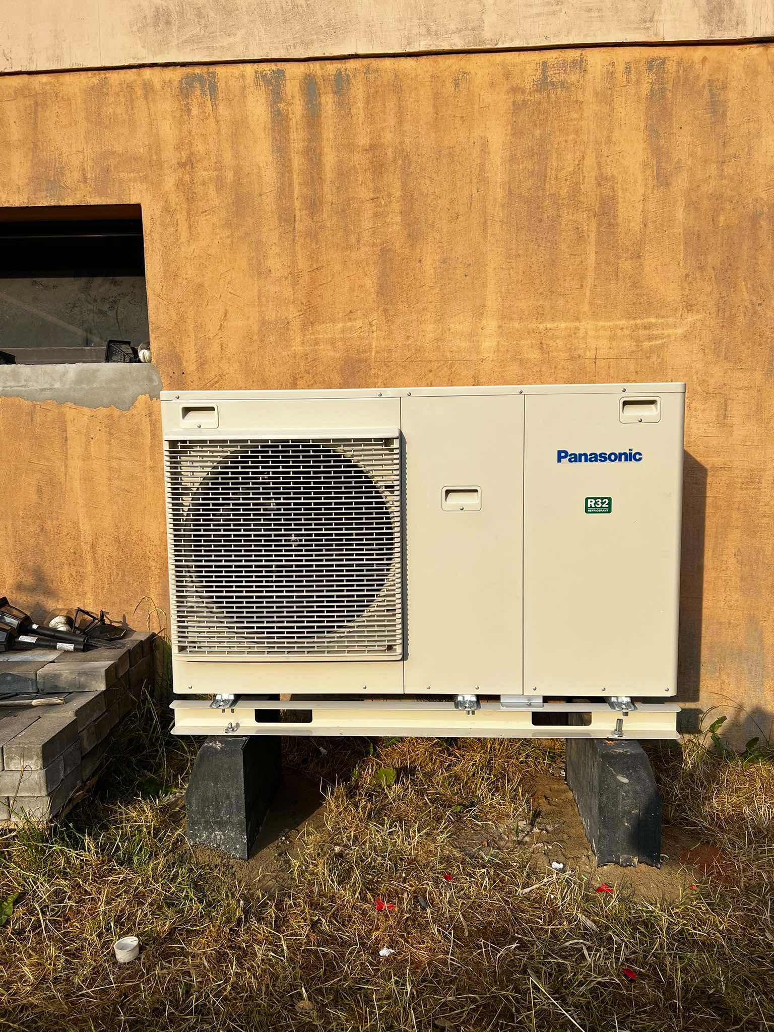 Kompaktowa pompa ciepła Panasonic R32 na betonowych podstawach przed ścianą o fakturze tynku rustykalnego, z widocznym fragmentem okna piwnicznego.