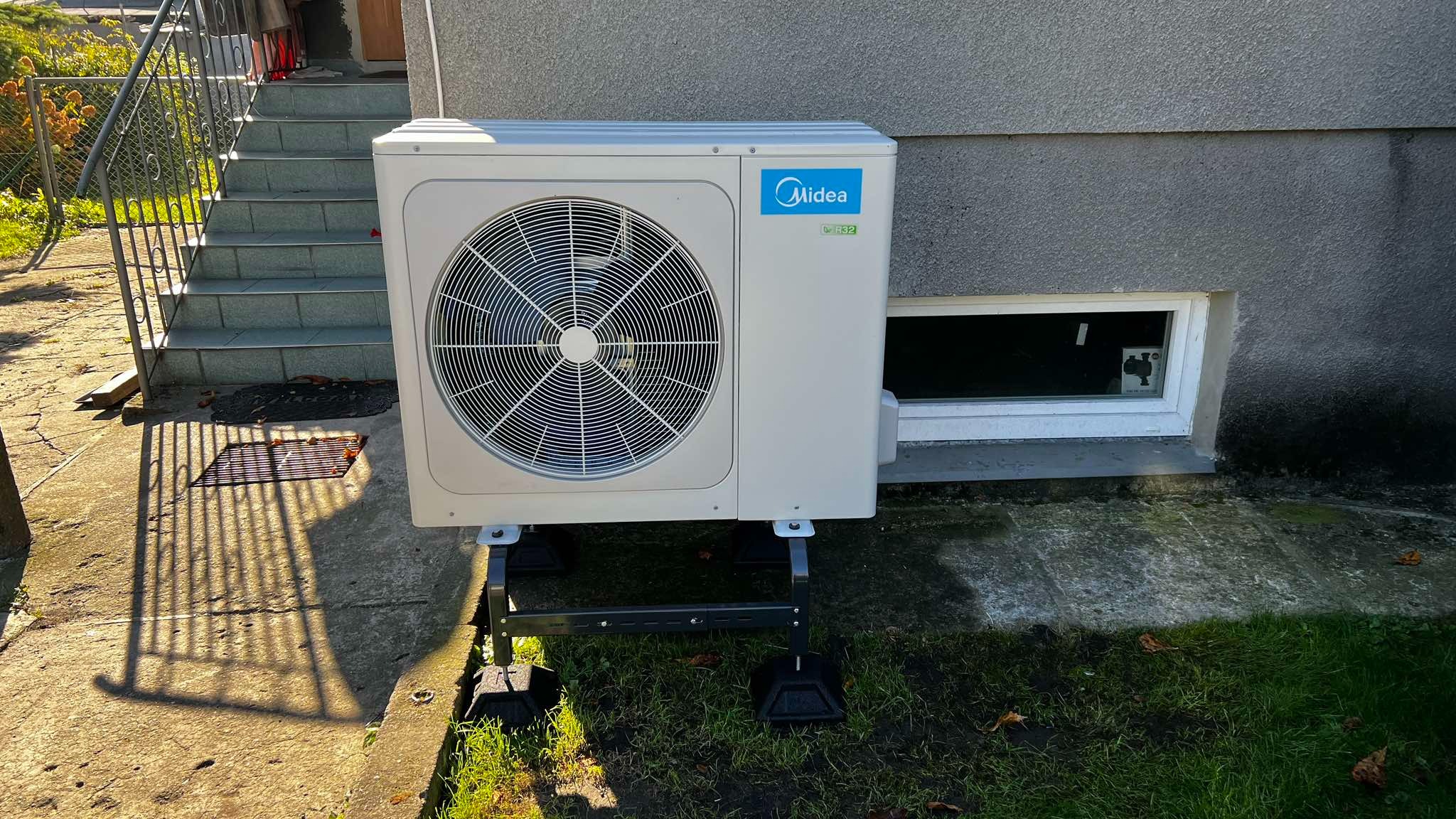 Biała jednostka zewnętrzna klimatyzacji Midea na czarnym stelażu, ustawiona na trawie obok betonowego budynku z małym oknem piwnicznym i schodami wejściowymi.