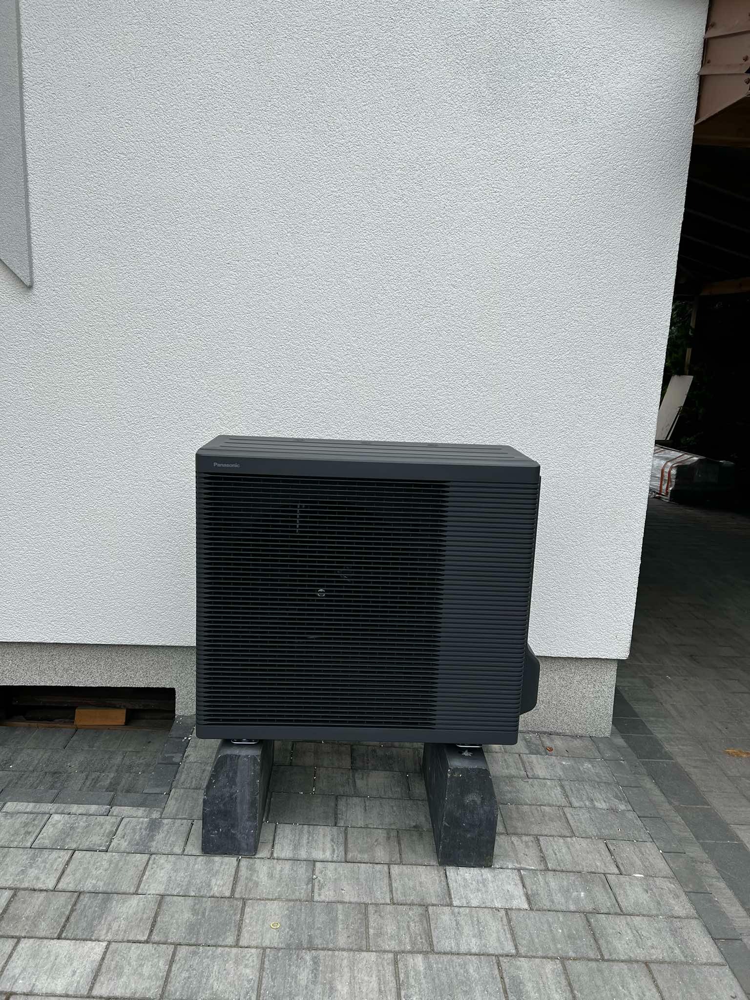 Czarna pompa ciepła Panasonic Aquarea, ustawiona na betonowych wspornikach przed białą ścianą budynku. Widoczna kostka brukowa i fragment dachu.