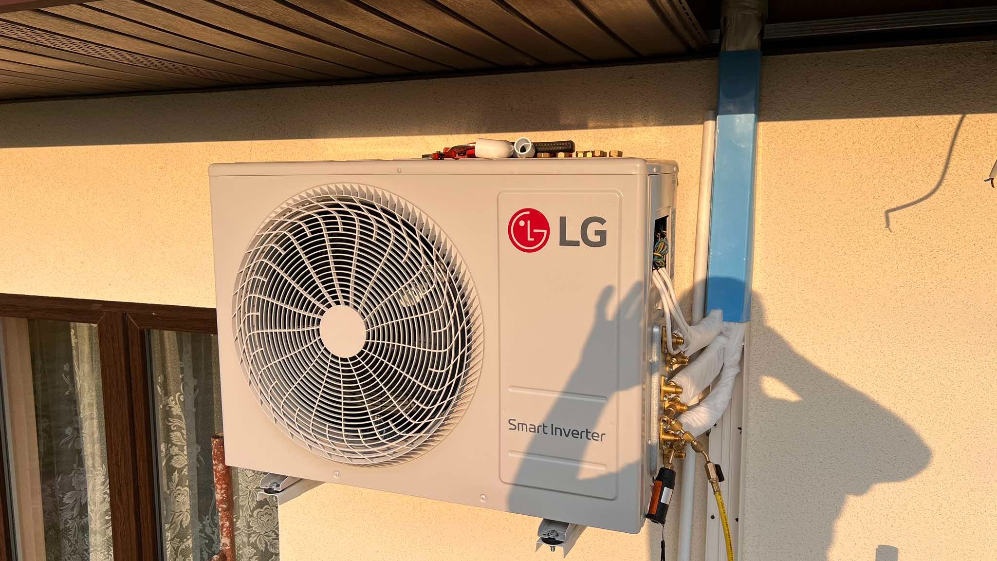 Zamontowana jednostka zewnętrzna klimatyzacji LG Smart Inverter na ścianie budynku, z widocznymi przewodami i narzędziami na górze obudowy. Ciepłe światło.