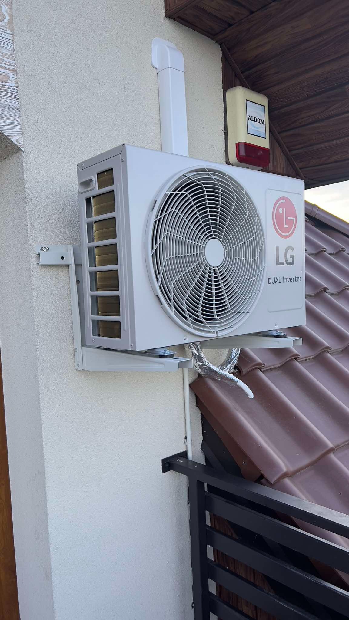 Biała jednostka zewnętrzna klimatyzacji LG Dual Inverter zamontowana na ścianie budynku z brązowym dachem i czarną balustradą w tle, widoczny przewód chłodniczy.