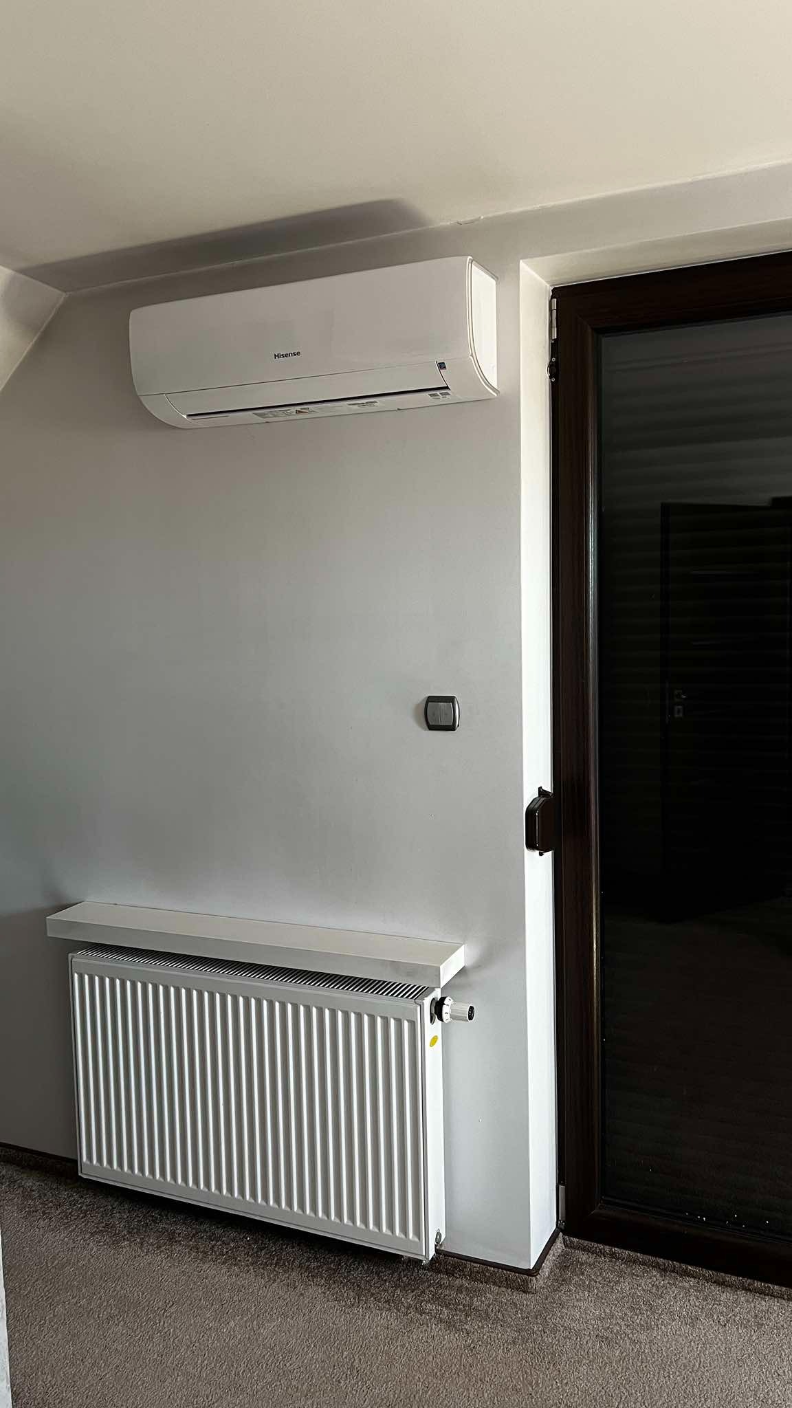 Biały klimatyzator Hisense nad grzejnikiem w Ujeździe, zamontowany na szarej ścianie obok ciemnych drzwi. Widoczny fragment podłogi.