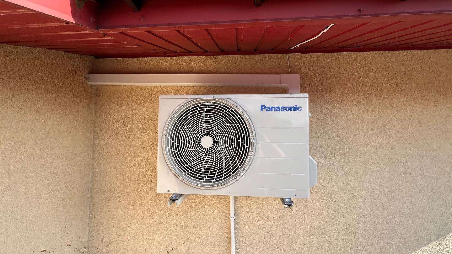 Zamontowana klimatyzacja Panasonic na beżowej ścianie pod czerwonym okapem. Widoczna biała rura instalacyjna i wsporniki montażowe. Ściana z fakturą baranka.
