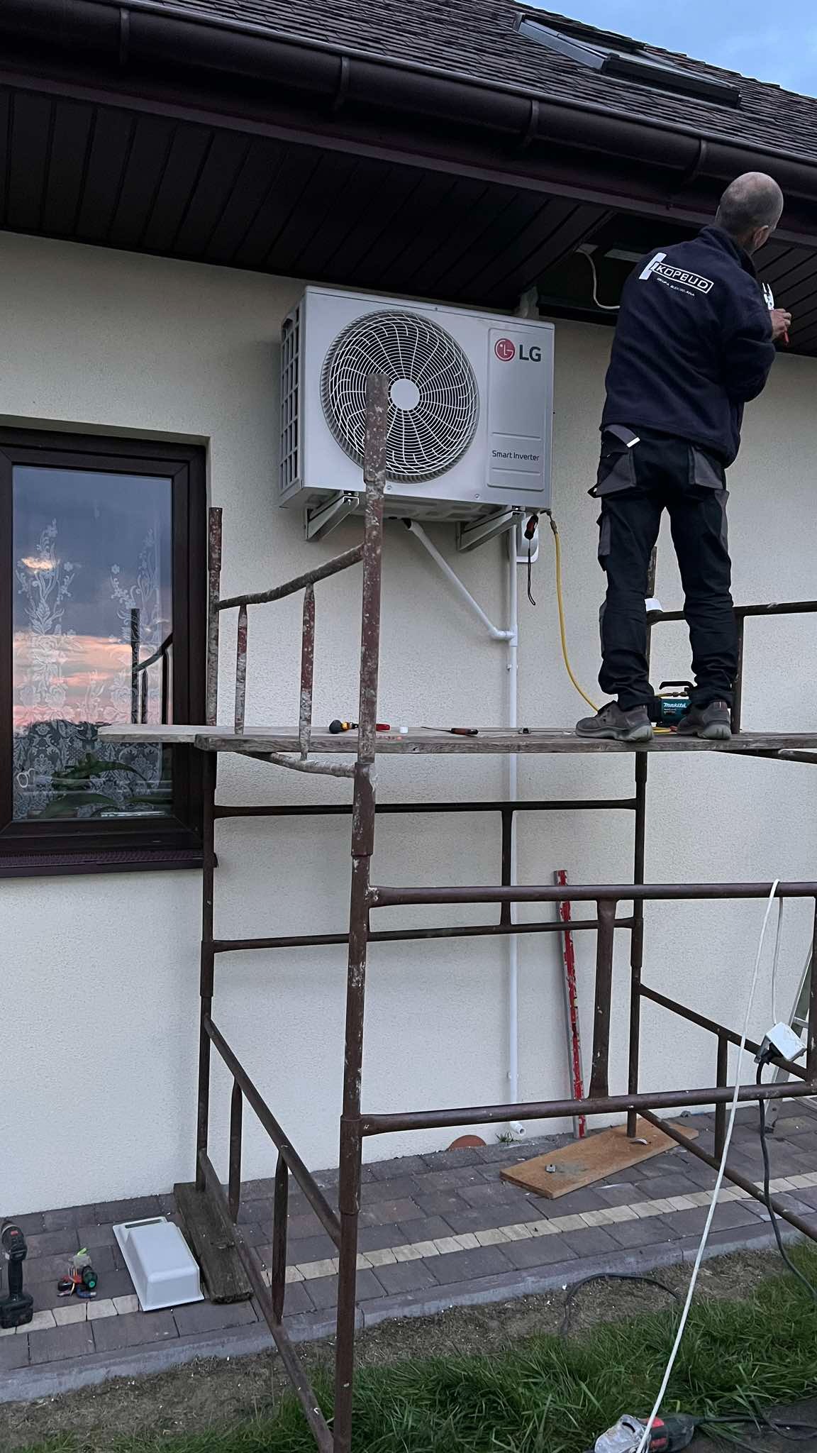 Montaż klimatyzacji LG Smart Inverter na elewacji domu. Instalator na rusztowaniu, narzędzia w pobliżu. Widoczne okno z odbiciem zachodu słońca.