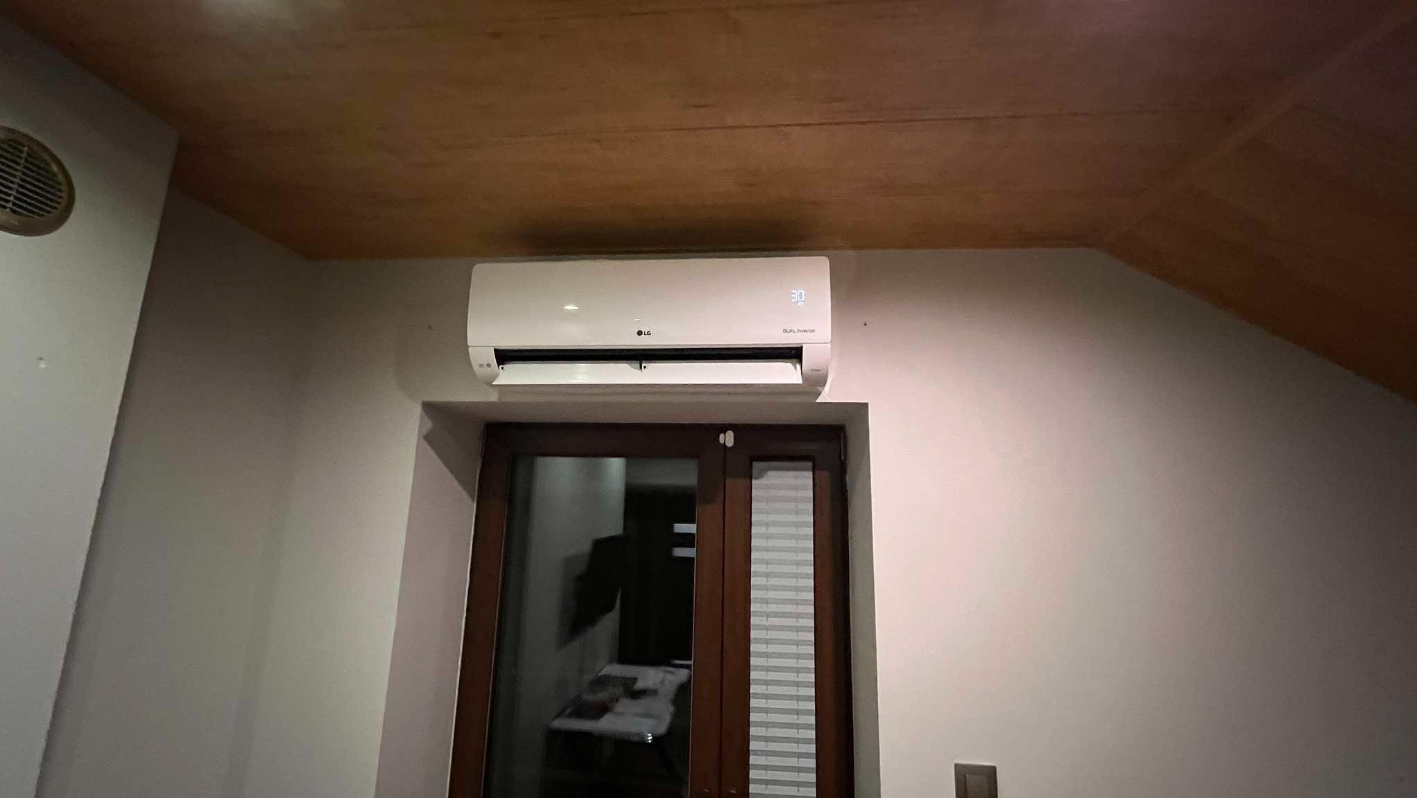 Biały klimatyzator LG zamontowany nad ciemnymi, przeszklonymi drzwiami balkonowymi na poddaszu, z widocznym wyświetlaczem temperatury 30 stopni.