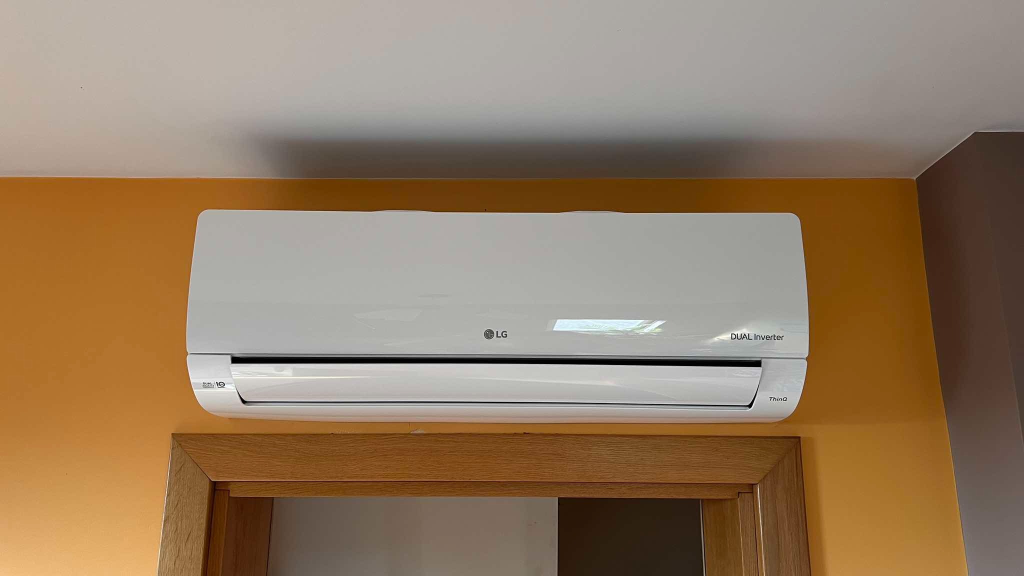 Biały klimatyzator LG Dual Inverter na pomarańczowej ścianie, nad drewnianą futryną drzwi. Widok z dołu, lekko z prawej strony. Widoczny fragment sufitu i brązowej ściany.