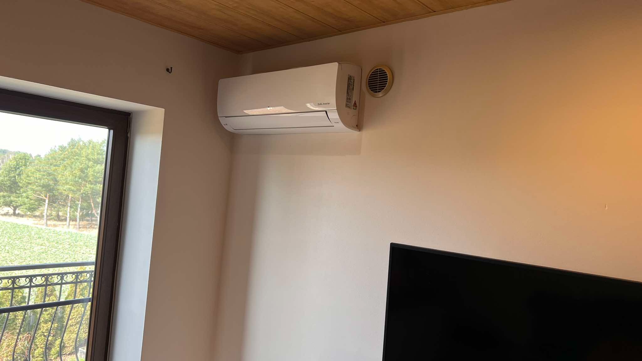 Biały klimatyzator ścienny z okrągłą kratką wentylacyjną obok, zamontowany w jasnym wnętrzu z drewnianym sufitem i widokiem na zieleń przez okno.