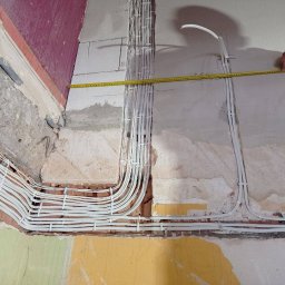 EK-MONTAZ - Instalacja elektryczna w trakcie montażu: białe przewody ułożone w korytach w surowej ścianie, pomiar odległości taśmą mierniczą.