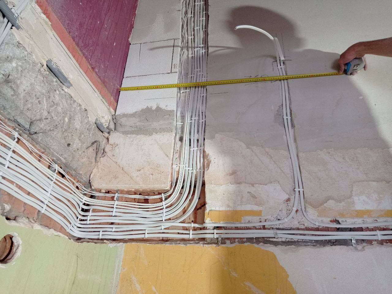 Instalacja elektryczna w trakcie montażu: białe przewody ułożone w korytach w surowej ścianie, pomiar odległości taśmą mierniczą.