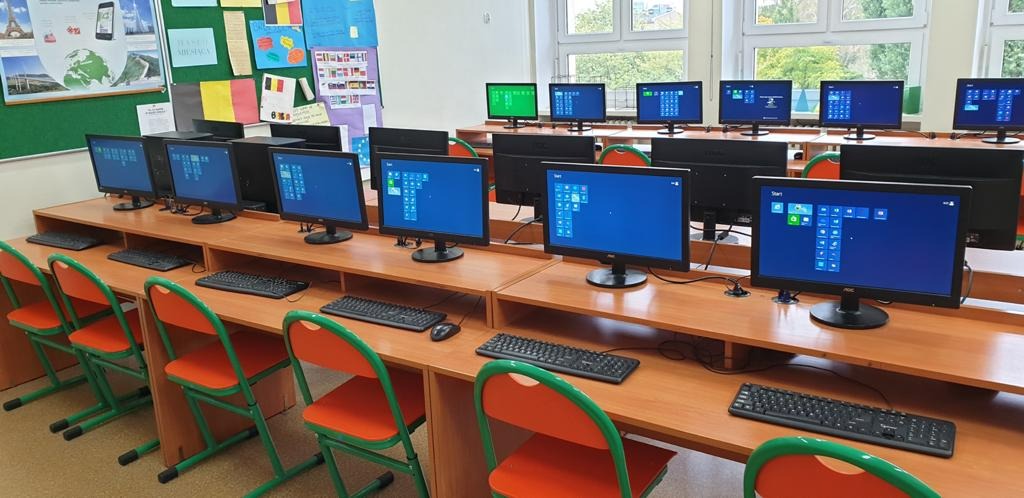 Sala komputerowa z rzędem biurek, na których stoją monitory i klawiatury. Monitory wyświetlają ekran startowy systemu Windows. W tle widoczne okna i tablica korkowa z notatkami.