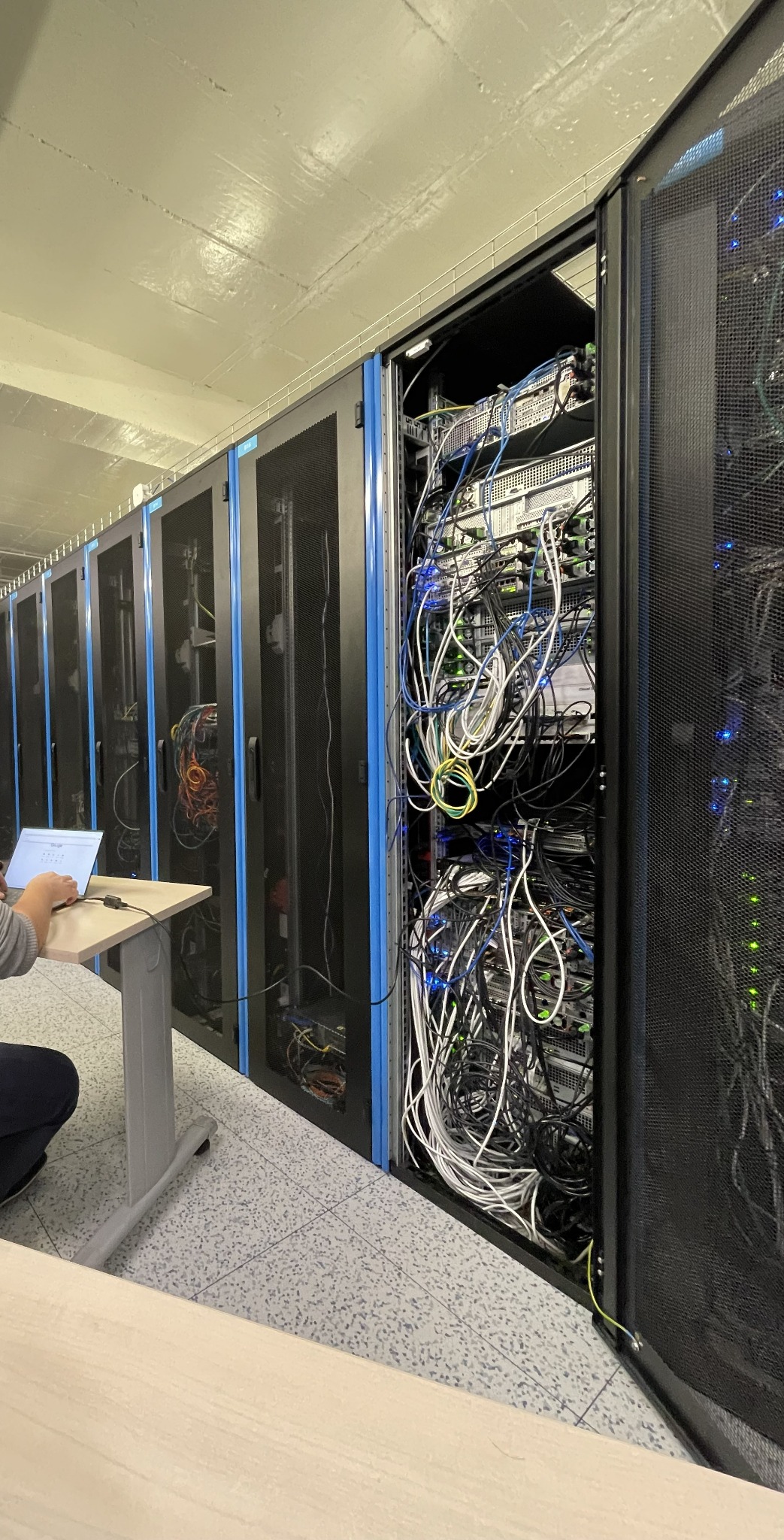 Rząd szaf serwerowych z niebieskimi akcentami, otwarte drzwi jednej szafy ukazują gąszcz kabli, pracownik IT z laptopem przy biurku w lewym dolnym rogu.
