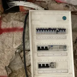 Otwarta skrzynka elektryczna z bezpiecznikami na starej, pomalowanej na czerwono ścianie, z widocznymi kablami wchodzącymi do ściany.