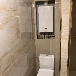 Nowoczesna łazienka z beżowymi marmurkowymi płytkami, wiszącą toaletą i białym piecem gazowym w zabudowie.