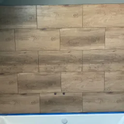 Ułożone płytki imitujące drewno na ścianie w łazience, widoczne przyłącza hydrauliczne, zabezpieczona wanna.