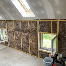 Poddasze w trakcie izolacji termicznej: wełna mineralna między metalowymi profilami, widoczne okna dachowe i narzędzia budowlane.