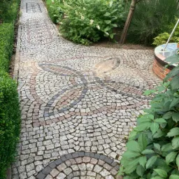 Kamienna ścieżka w ogrodzie z dekoracyjnym wzorem z kostki brukowej w odcieniach szarości, różu i czerni, otoczona zielenią krzewów i drzew.