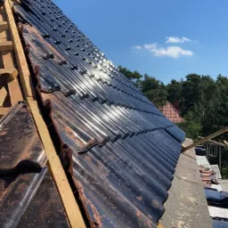 Układanie czarnej dachówki ceramicznej na drewnianej konstrukcji więźby dachowej, widok z dołu na tle błękitnego nieba i zieleni drzew.