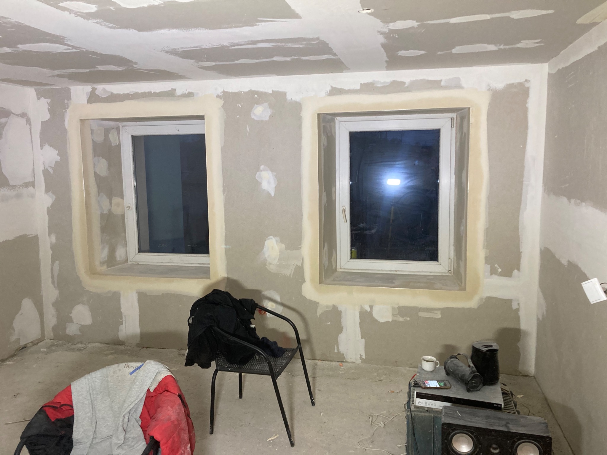 Pokój w trakcie prac wykończeniowych: ściany i sufit pokryte gładzią szpachlową, widoczne dwa okna z obrobionymi ościeżami, na podłodze narzędzia i ubrania.