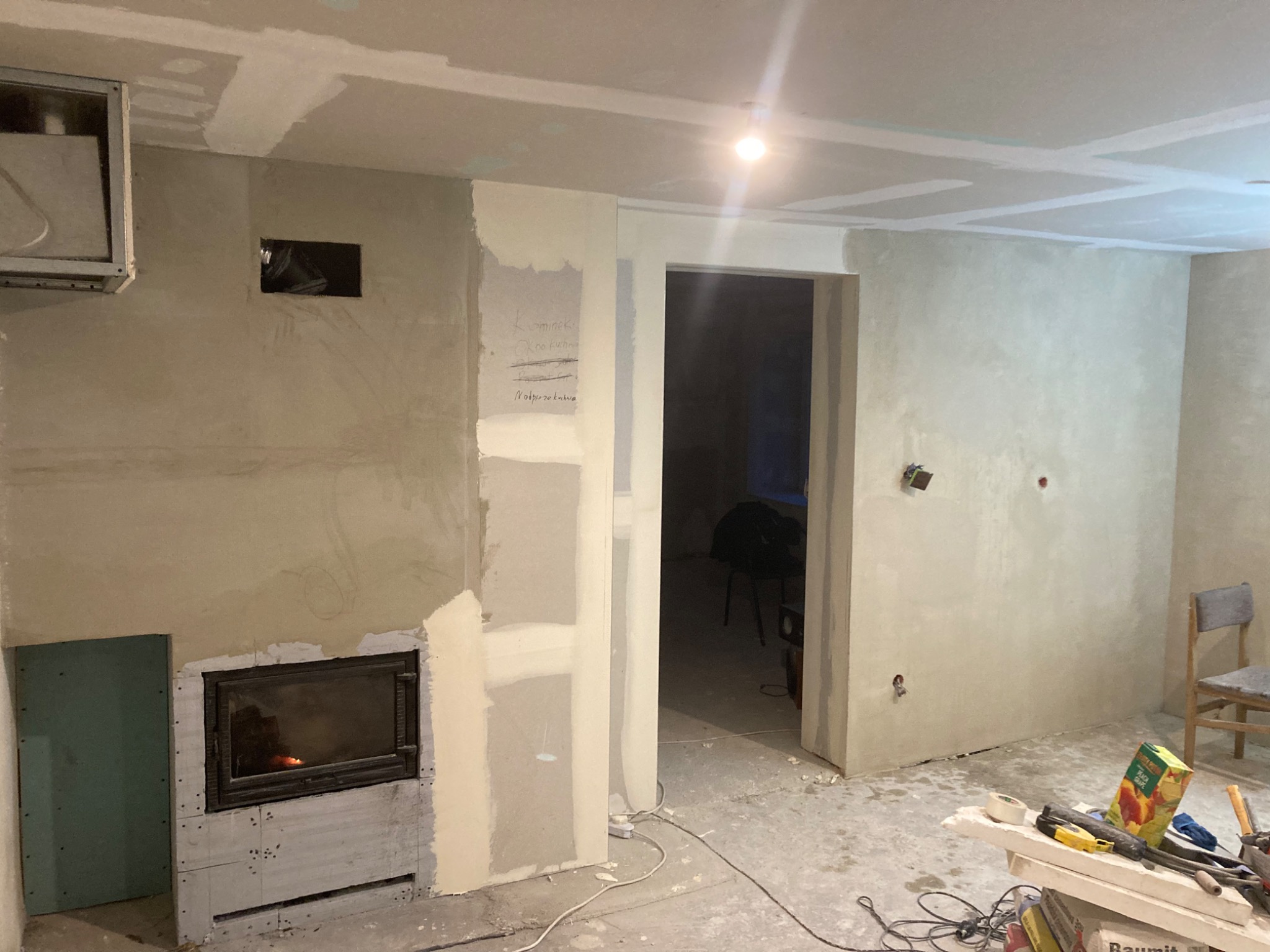 Wnętrze w trakcie prac wykończeniowych: widoczny wkład kominkowy, częściowo obłożony płytami, ściany przygotowane do malowania, sufit z zamontowanymi płytami, notatki na ścianie, narzędzia...