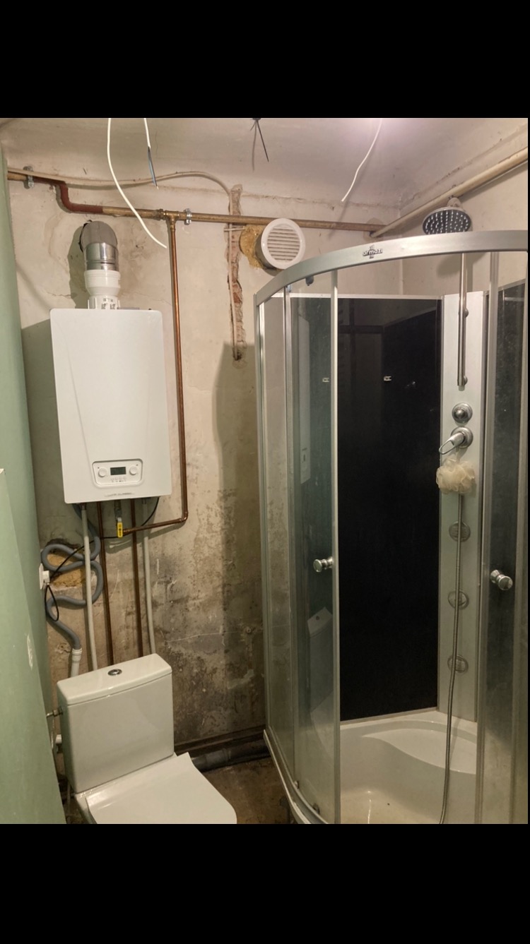Niedokończona łazienka z kotłem gazowym, toaletą i kabiną prysznicową z hydromasażem na tle surowej ściany z widocznymi rurami.