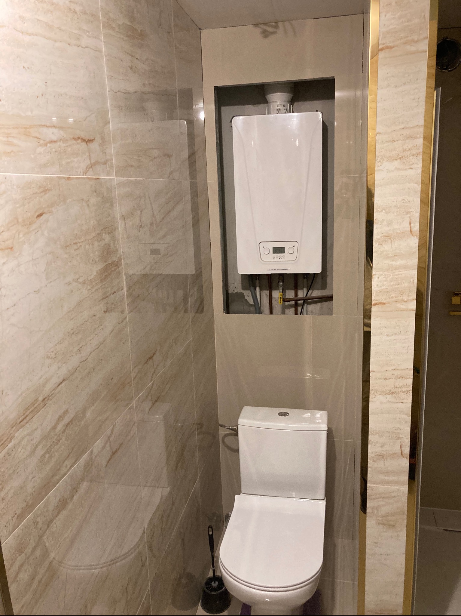 Nowoczesna łazienka z beżowymi marmurkowymi płytkami, wiszącą toaletą i białym piecem gazowym w zabudowie.