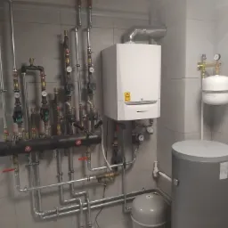 Nowoczesna kotłownia z białym piecem gazowym, szarym zbiornikiem na wodę i rozbudowaną instalacją hydrauliczną z licznymi zaworami i manometrami na tle szarych płytek.