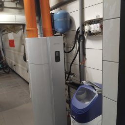 KAM-INSTAl - Biały, pionowy bojler Atlantic z pomarańczowymi rurami, niebieskim zbiornikiem wyrównawczym i zmiękczaczem wody w pomieszczeniu z białymi płytkami. Widoczna instalacja elektryczna.