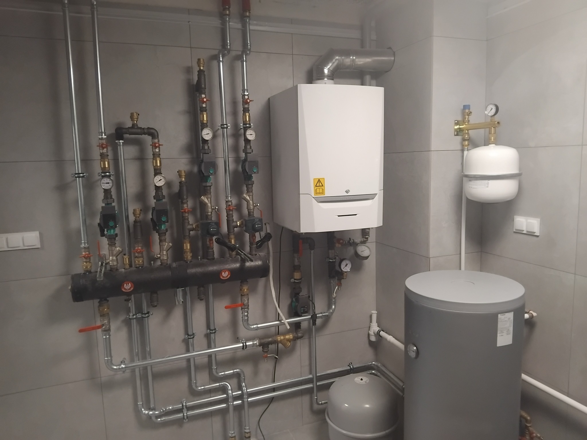 Nowoczesna kotłownia z białym piecem gazowym, szarym zbiornikiem na wodę i rozbudowaną instalacją hydrauliczną z licznymi zaworami i manometrami na tle szarych płytek.