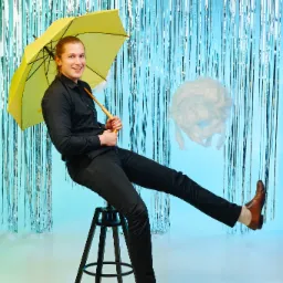 Uśmiechnięty mężczyzna w czarnym stroju siedzi na stołku z żółtym parasolem na tle błyszczącej kurtyny i sztucznej chmury.