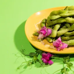 Żółty talerz pełen zielonych strąków edamame posypanych solą, ozdobiony różowymi kwiatami groszku pachnącego na jasnozielonym tle, w jasnym oświetleniu studyjnym.