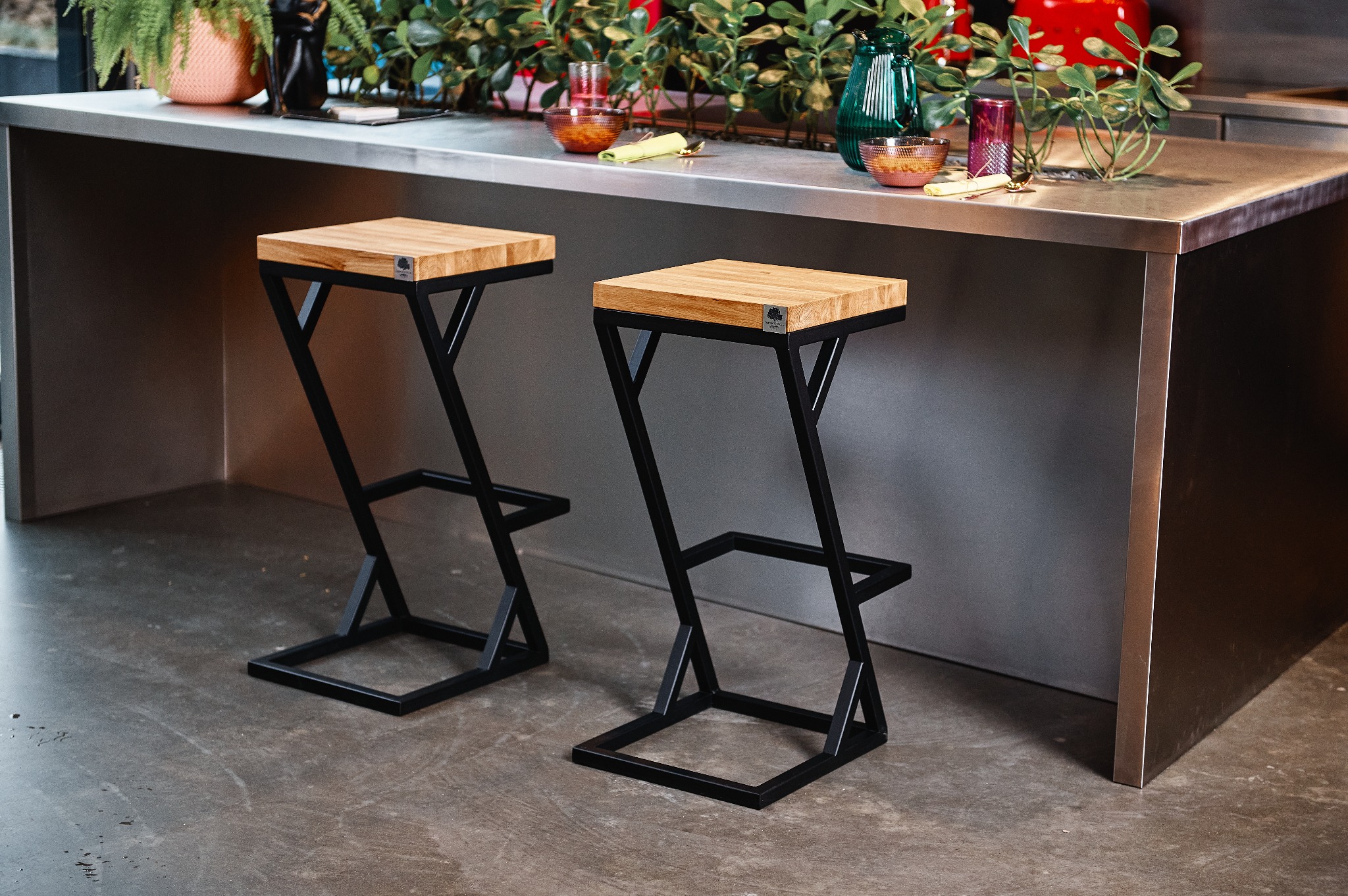 Dwa nowoczesne hokery z drewnianymi siedziskami i czarnymi metalowymi nogami w kształcie litery Z, ustawione przed metalowym blatem z dekoracjami roślinnymi i szklanymi naczyniami.