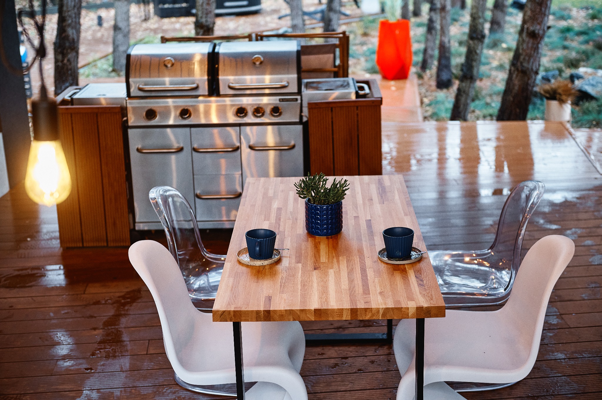 Zewnętrzna przestrzeń jadalna z drewnianym stołem, dwoma filiżankami, rośliną w niebieskiej doniczce, przezroczystymi krzesłami, grillem ze stali nierdzewnej w tle i żarówką na pierwszym planie.