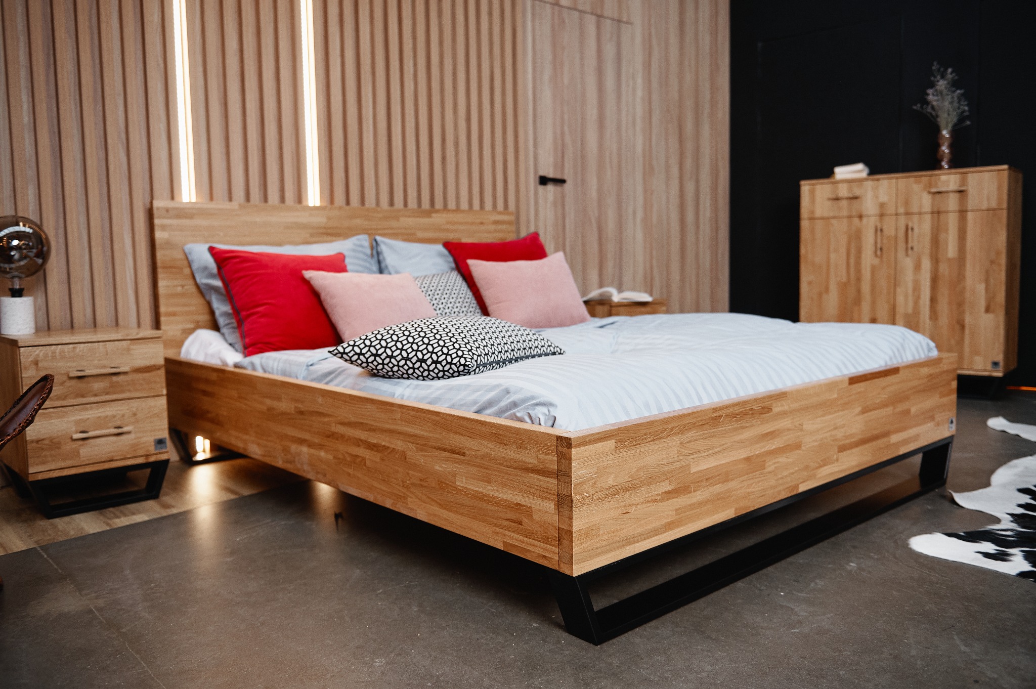 Drewniane łóżko z czarnymi nogami, czerwonymi i różowymi poduszkami oraz szafkami nocnymi w minimalistycznej sypialni.