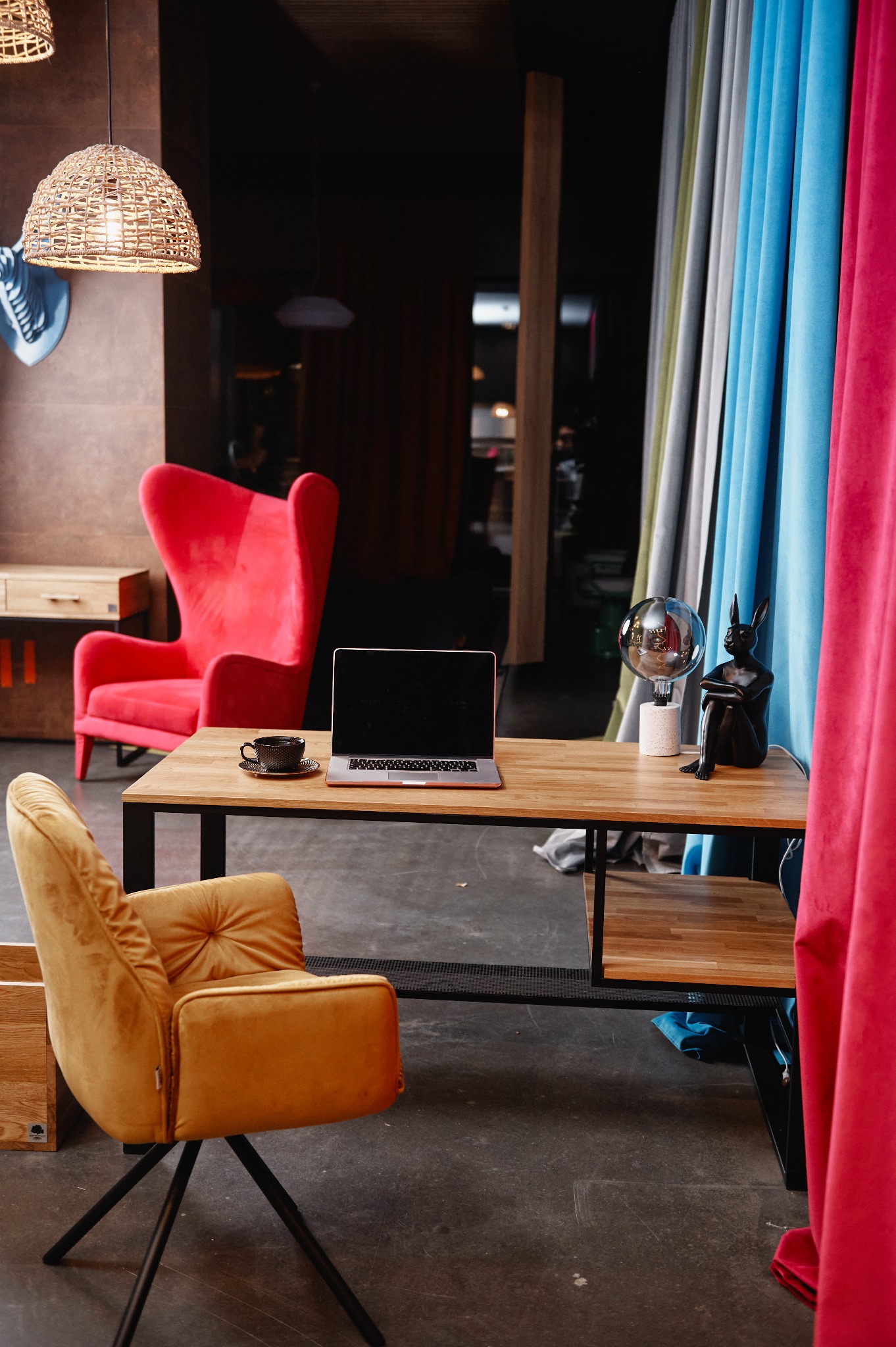 Nowoczesne biuro z drewnianym biurkiem, laptopem, filiżanką kawy, lampką w kształcie kuli i czarną figurką królika, z czerwonym i musztardowym fotelem w tle oraz zasłonami w odcieniach szarości...
