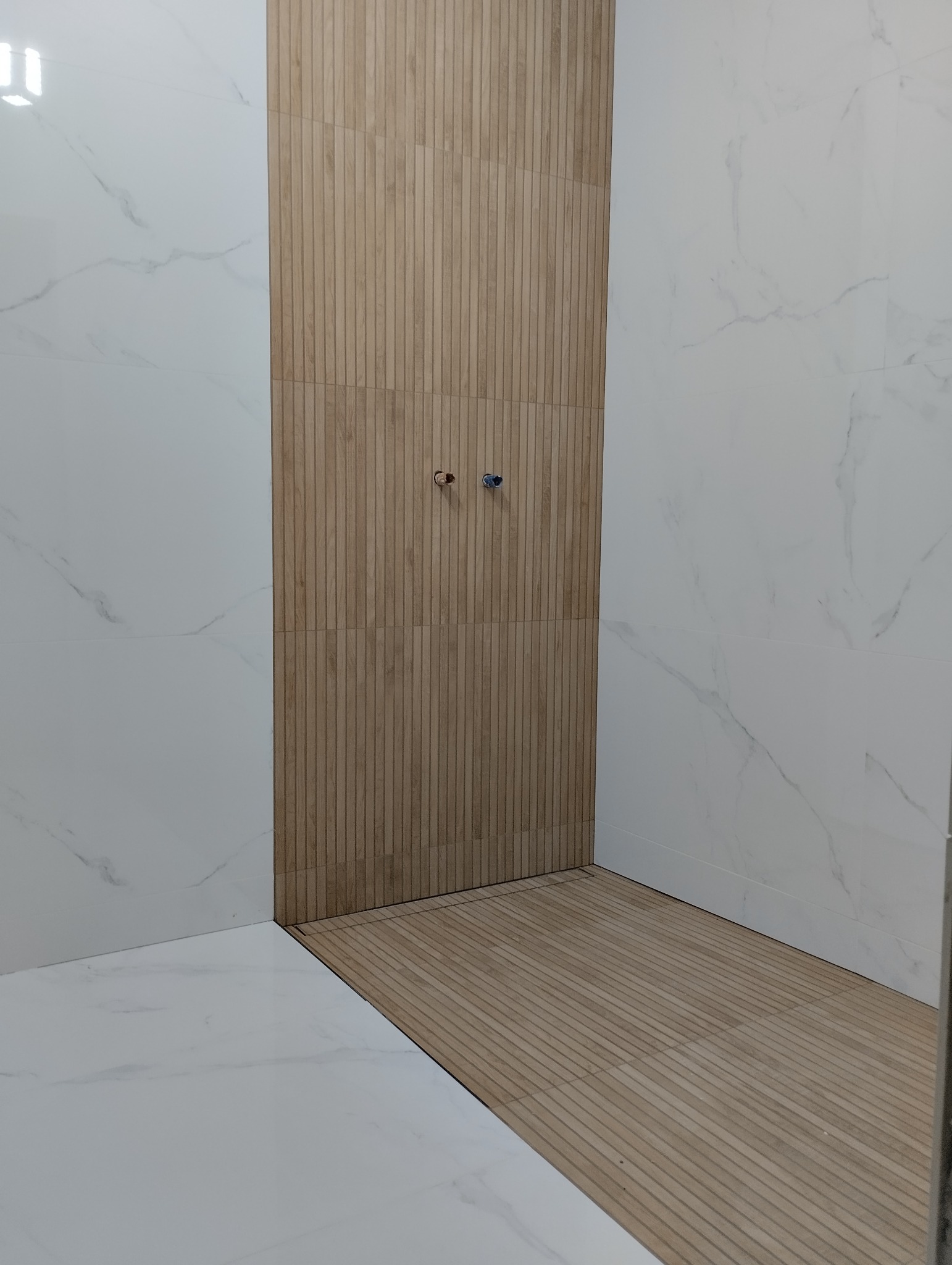 Nowoczesna łazienka w trakcie wykańczania, z białymi marmurkowymi płytkami na ścianach i drewnopodobnymi pionowymi płytkami w strefie prysznicowej z widocznymi przyłączami wodnymi.