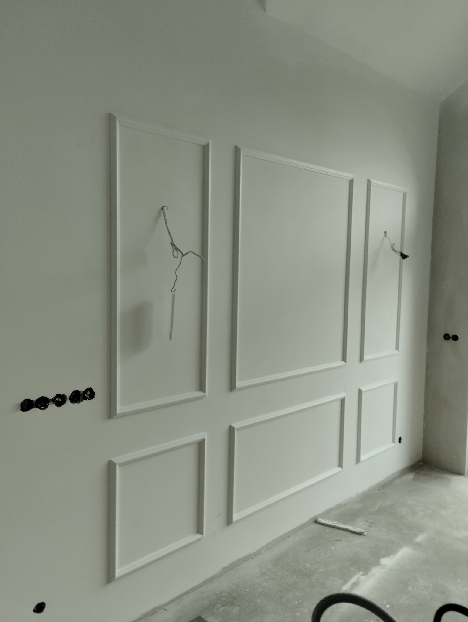 Ściana z dekoracyjnymi, białymi panelami w stylu klasycznym, z widocznymi przewodami i gniazdkami elektrycznymi w trakcie montażu.