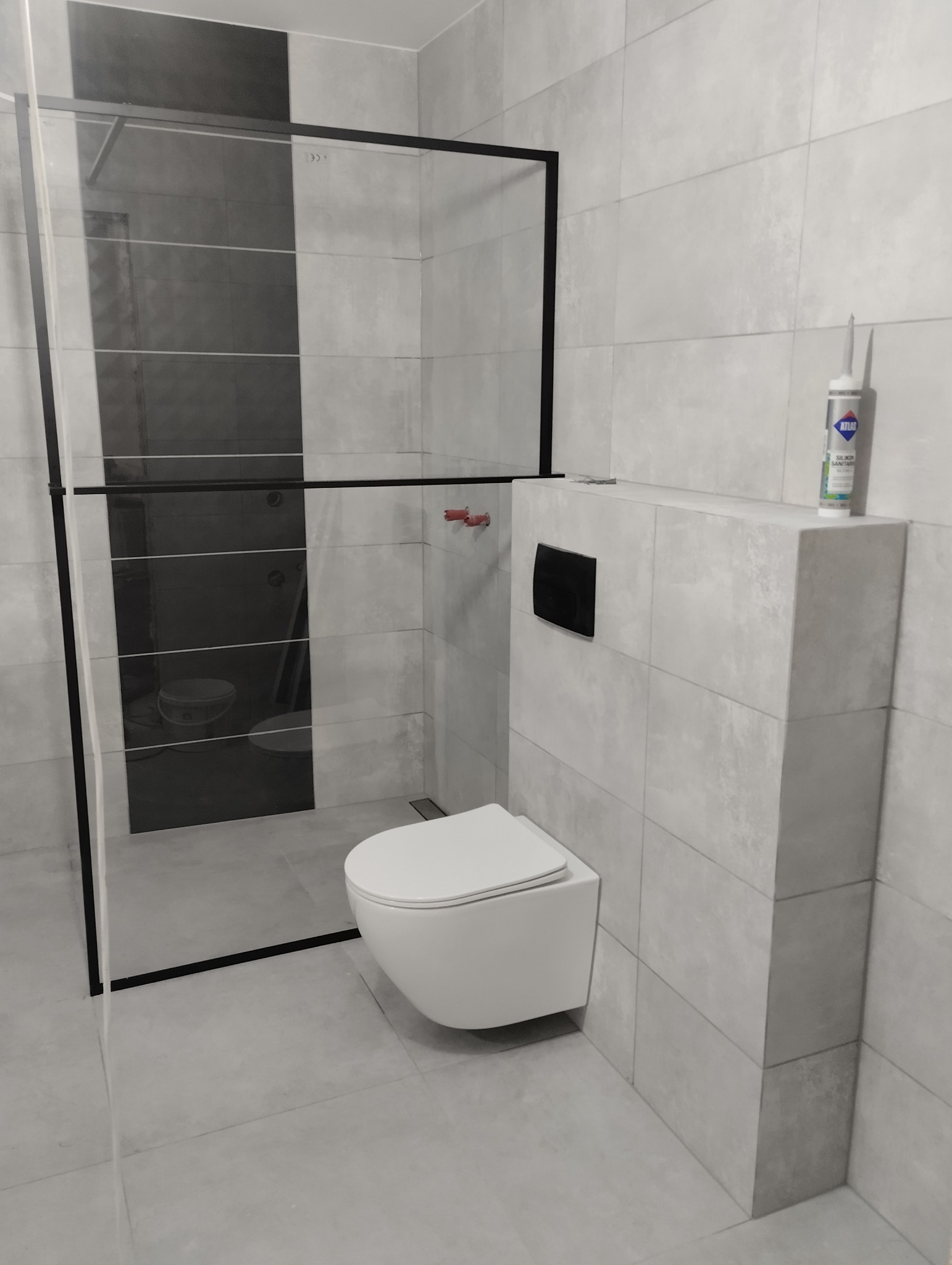 Nowoczesna łazienka w szarościach z kabiną prysznicową typu walk-in z czarnymi profilami i podwieszaną miską WC; widoczny silikon sanitarny na zabudowie stelaża.