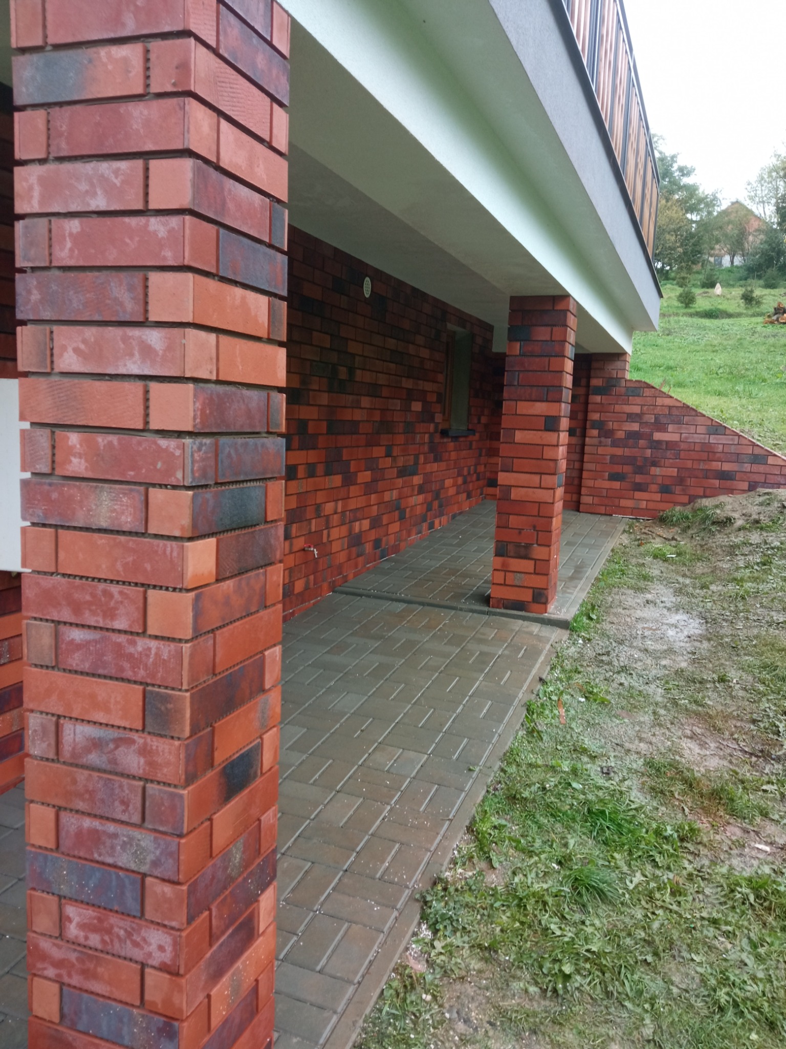 Wykończony taras z ceglanymi filarami i szarą kostką brukową, widok na zielony trawnik i fragment budynku z drewnianą balustradą.