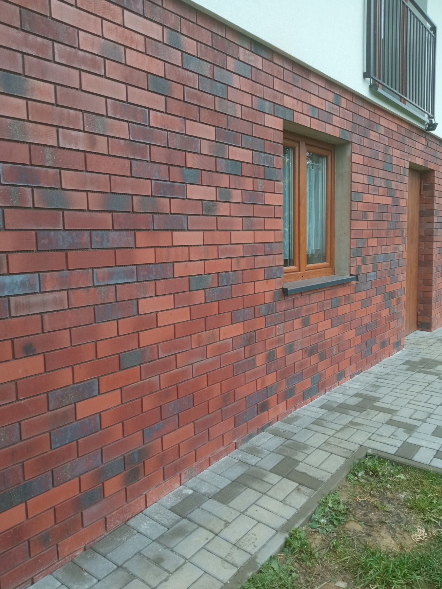 Elewacja budynku wykończona czerwoną cegłą klinkierową z ciemnymi przebarwieniami, widoczne okno z drewnianą ramą i fragment chodnika z kostki brukowej.