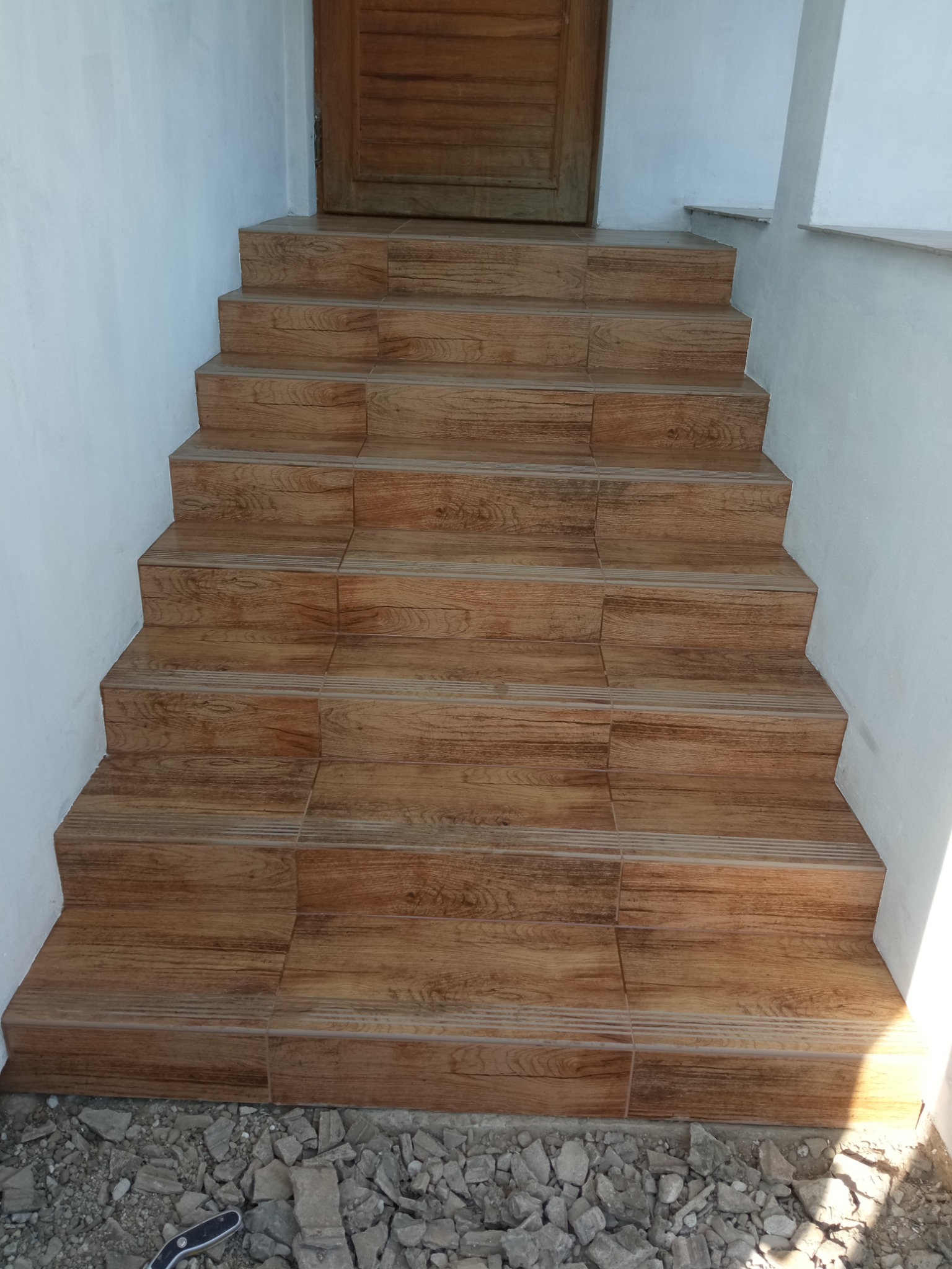 Wykończone schody wejściowe z brązowych płytek imitujących drewno, prowadzące do drewnianych drzwi. Widoczne białe ściany i gruz u podstawy schodów.