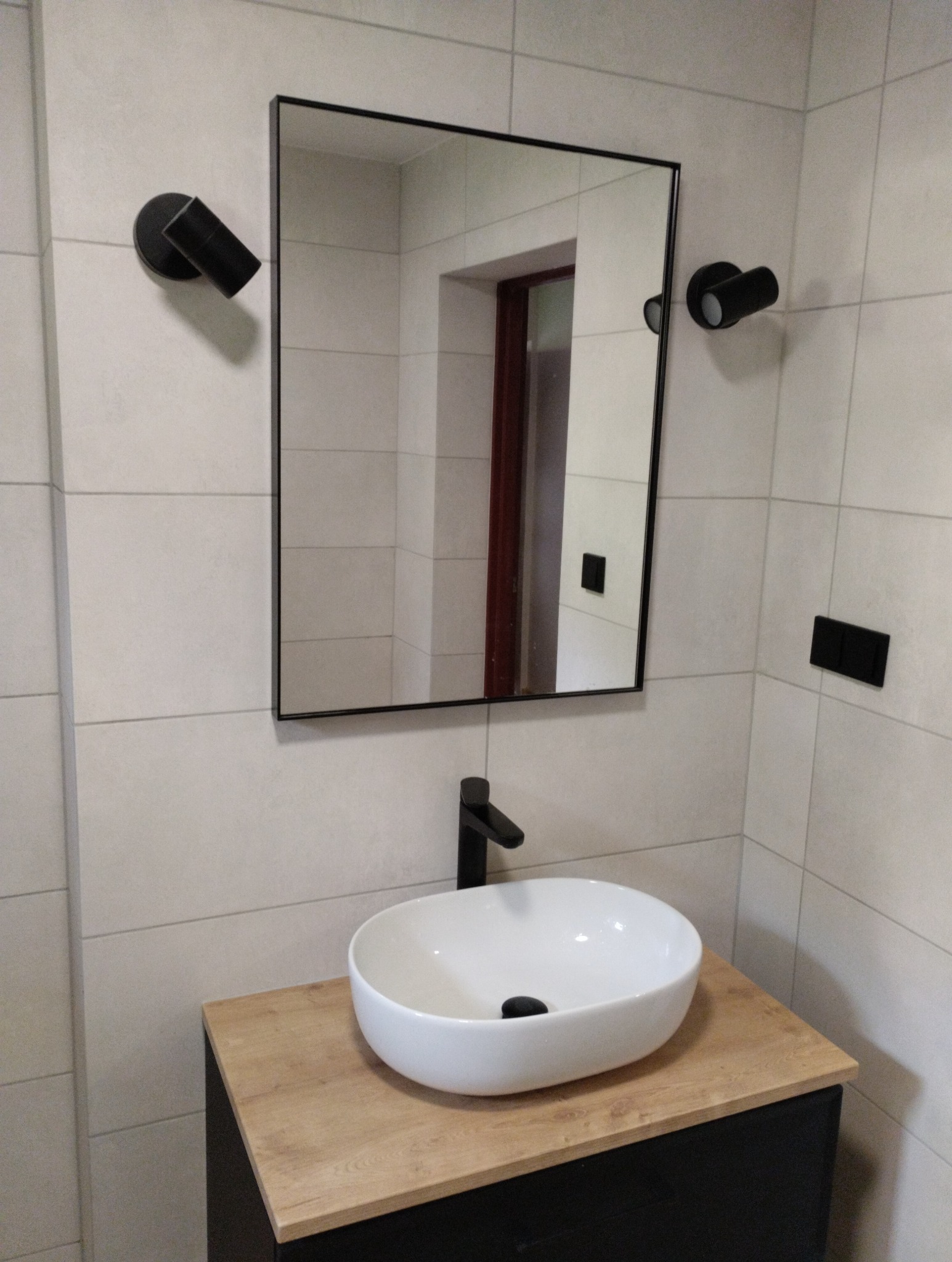 Nowoczesna umywalka nablatowa na szafce z blatem drewnopodobnym, czarna bateria i okrągłe lampy ścienne w wykończonej kafelkami łazience.