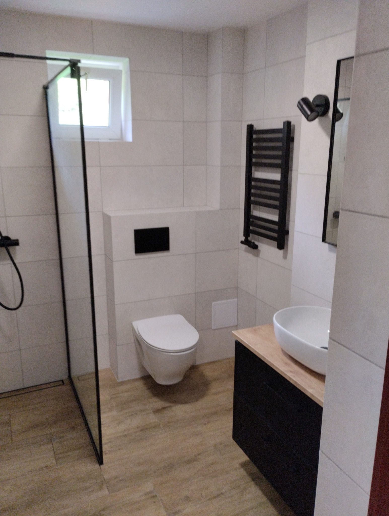 Nowoczesna łazienka z kabiną typu walk-in, podwieszaną toaletą i czarnym grzejnikiem drabinkowym. Umywalka nablatowa na szafce z drewnianym blatem.