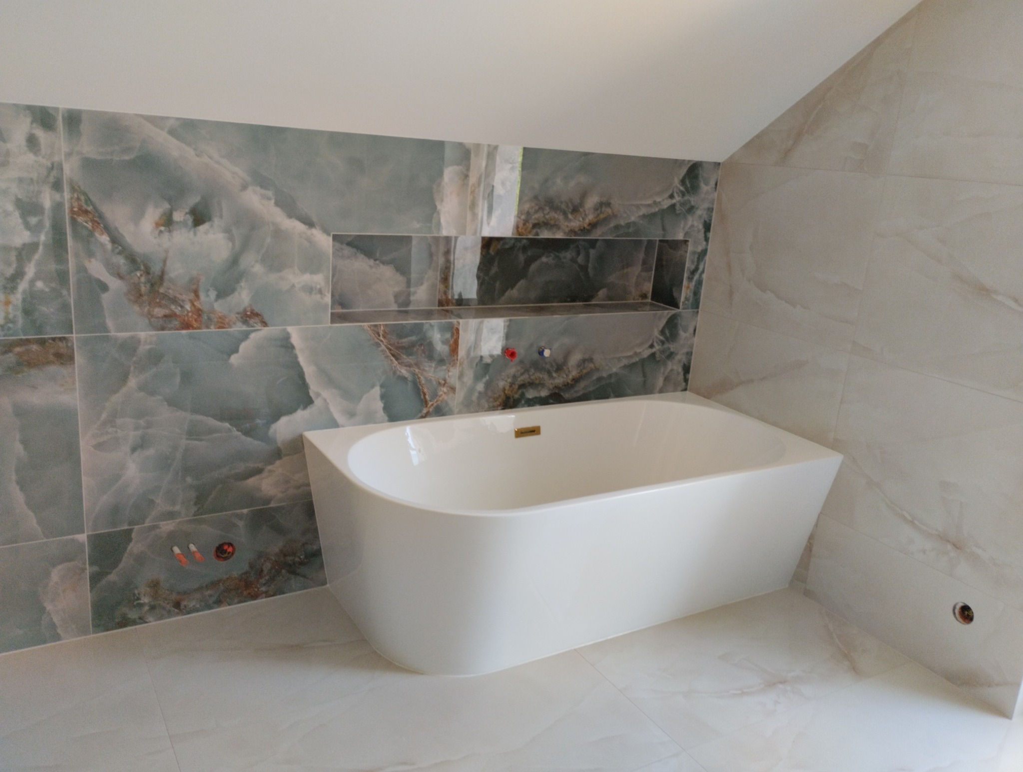 Biała wanna wolnostojąca w łazience z marmurowymi płytkami w odcieniach błękitu i beżu. Widoczne wnęki w ścianie wyłożone tym samym materiałem.