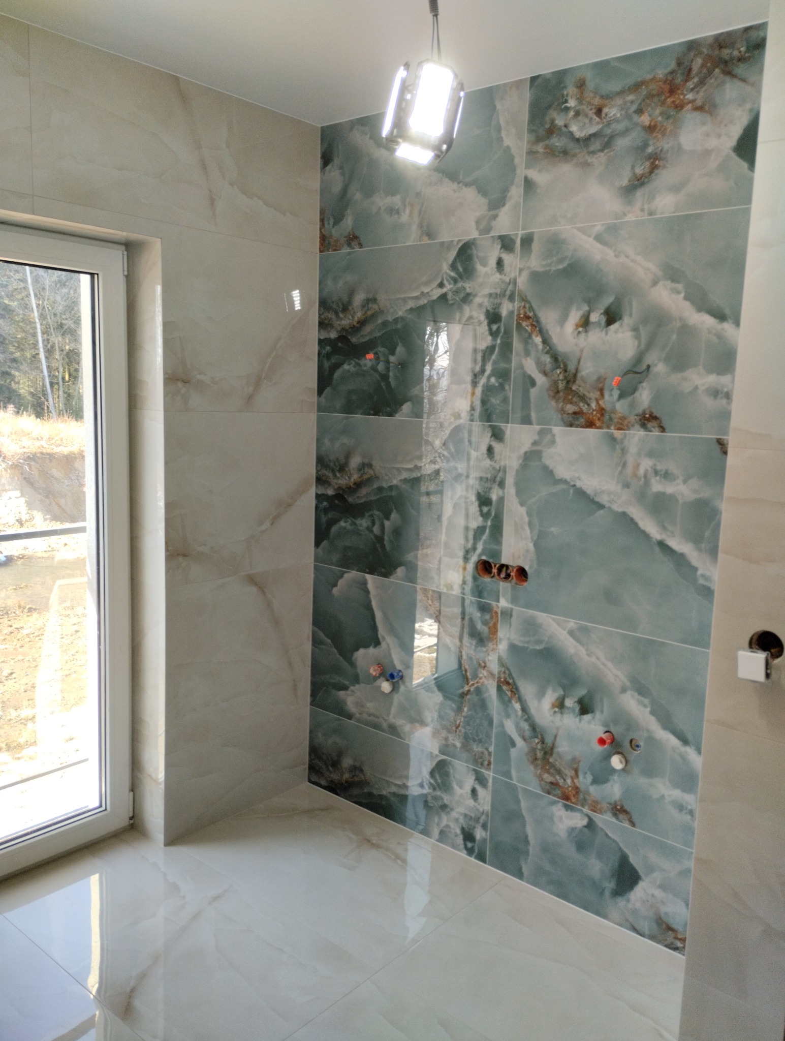 Wykończona łazienka z marmurowymi płytkami w odcieniach bieli i turkusu. Widoczne przygotowane punkty instalacyjne. Nowe okno z widokiem na zieleń.