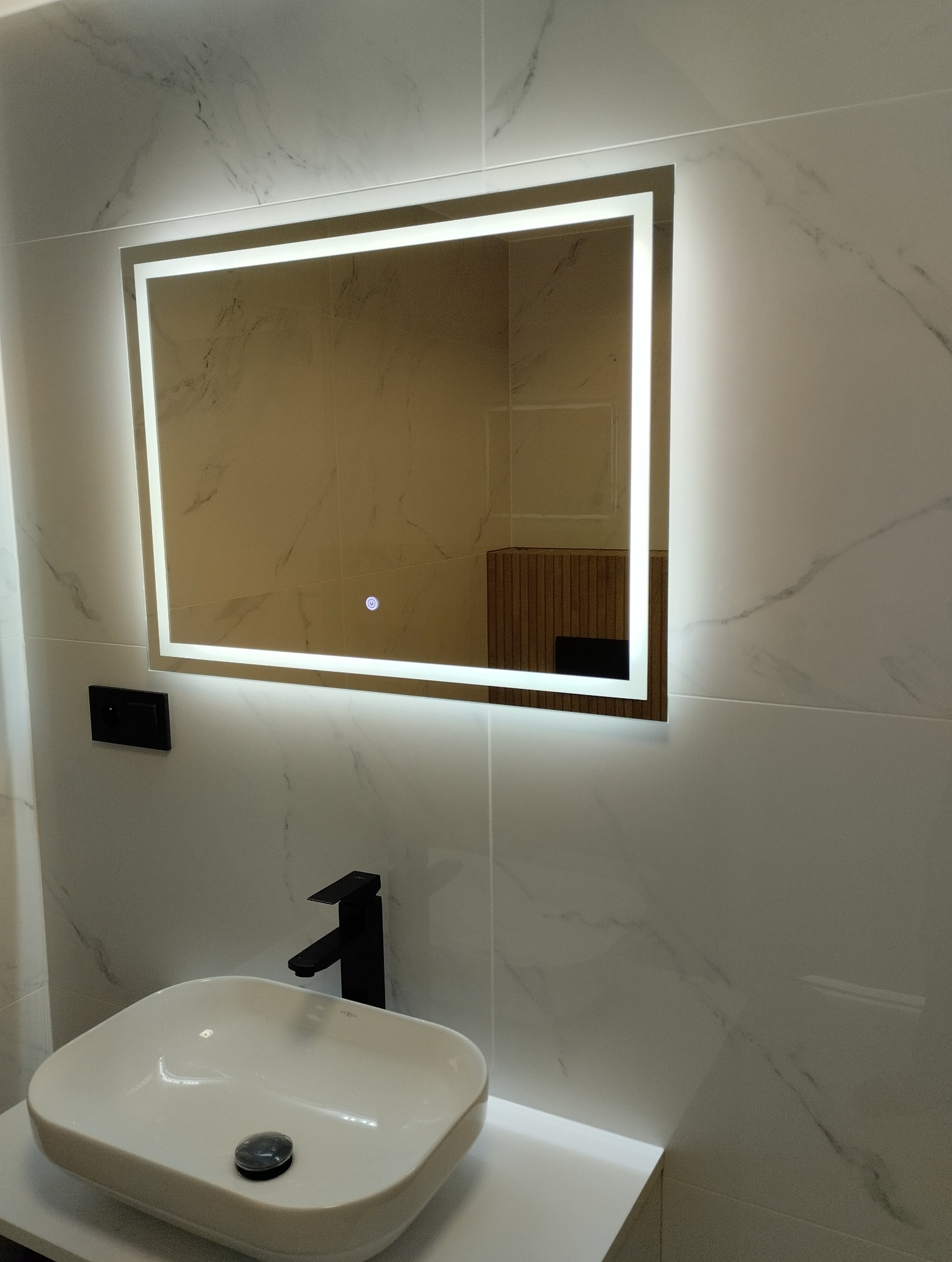 Nowoczesna łazienka z podświetlanym lustrem, białą umywalką nablatową i czarną armaturą na tle marmurowych płytek. Minimalistyczny design.